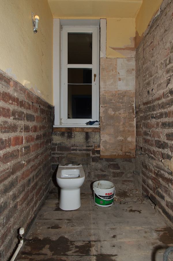 Een smalle badkamer tijdens de renovatie: bakstenen muren zonder tegels, een toilet en een emmer op de ruwe vloer