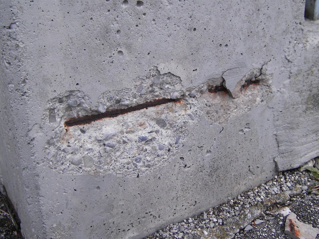 Een betonnen muur waar een stuk beton is afgesprongen en het roestige wapeningsijzer bloot ligt